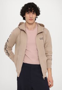 Hanorac bej cu fermoar și glugă cu şnur; are un logo negru și text pe mâneca stângă; textură moale, asociat cu un tricou roz deschis și pantaloni închiși.