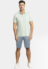 Camisa polo de color verde menta claro, de mangas cortas con cuello, combinada con pantalones cortos azules claros y zapatillas blancas. Diseño simple y casual.