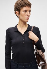 Mulher com cabelo curto, vestindo uma camisola de mangas longas preta em tecido canelado, segurando uma mala castanha com um lenço sobre um ombro.