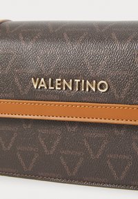 Καφέ δερμάτινη τσάντα χειρός με ανάγλυφο μοτίβο λογότυπου, διαθέτοντας χρυσό λογότυπο και καφέ δερμάτινη λεπτομέρεια στη λαβή. Υφή επιφάνειας.