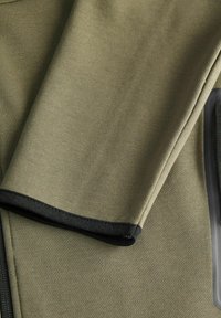 Tessuto di felpa verde oliva con una texture liscia. Presenta un orlo nero e dettagli con cerniera, enfatizzando elementi di design sportivo.