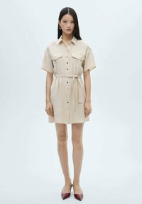 Abito camicia beige con maniche corte, chiusura a bottoni frontale, due tasche sul petto, vita cintrata con una cintura e una texture liscia. Indossato con tacchi a punta rossi.