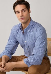 Man in een blauw geruit overhemd en een bruine broek, zittend op een licht houten stoel, kijkt iets opzij met zijn handen gevouwen.
