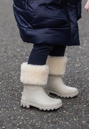 Kind draagt marineblauwe broek en marineblauwe jas met beige winterlaarzen gevoerd met dikke crèmekleurige bont, staand op asfalt.