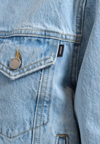 Chaqueta de mezclilla azul claro con cierre frontal de botones, costuras amarillas contrastantes y una etiqueta negra "DR. DENIM" en el bolsillo.