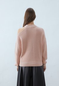 Pull tricoté rose clair avec une épaule dénudée, texture côtelée et manches longues, associé à une jupe plissée noire.