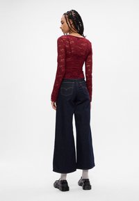 Top de renda burgundy de mangas longas combinado com calças de ganga escuras de perna larga. Apresenta padrões florais intrincados e uma silhueta ajustada.