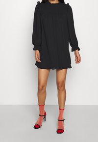 Robe noire à manches longues avec des accents froncés aux épaules et au col, en tissu texturé. Portée avec des chaussettes transparentes à paillettes rouges et des escarpins noirs.