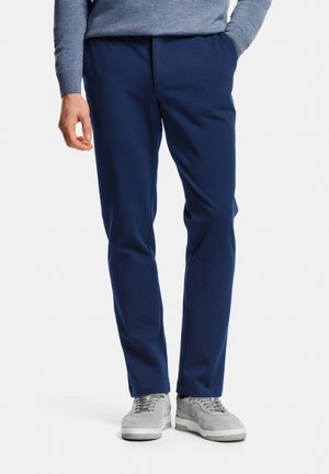 Man wearing navy blue slim-fit trousers, grey sneakers, and a light blue sweater with one hand in pocket, standing on white background.