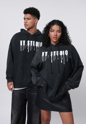 Svarta hoodies i en avslappnad passform med vit droppande text, kombinerat med svarta denimbyxor och en läderminikjol, på en neutral bakgrund.