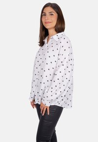 White button-up shirt with black heart pattern, flared long sleeves, and a curved hem. Paired with black fitted pants.