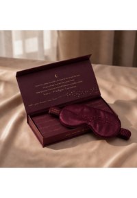 Dunkellila Seiden-Augenmaske mit dekorativen Perlen liegt auf einer offenen passenden Schachtel mit goldenem Text, auf einer beigen Satinoberfläche.
