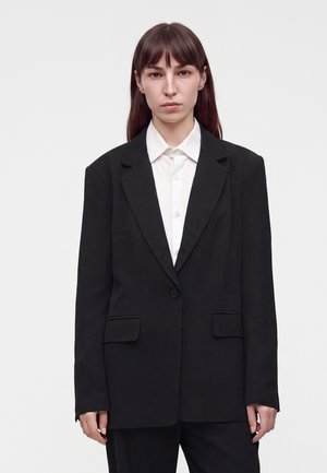 Vrouw met steil bruin haar draagt een zwarte oversized blazer over een witte blouse met knopen, staand tegen een effen lichte achtergrond.
