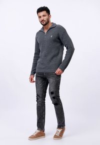 Maglione grigio a coste con collo con zip, abbinato a jeans neri strappati e sneakers color camoscio. Logo evidente sul petto.