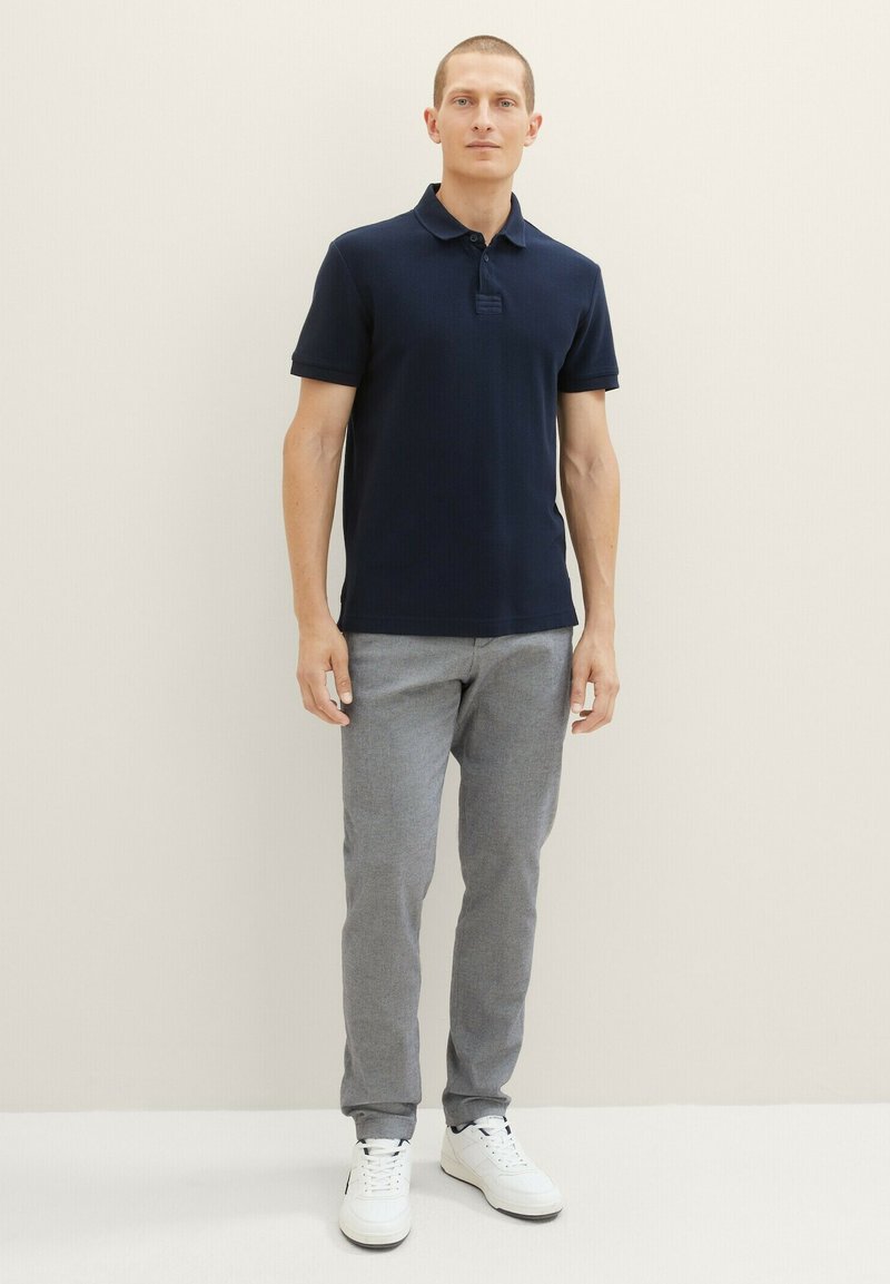 Marineblauwe polo met korte mouwen, gemaakt van een gestructureerde stof. Draagt het met lichtgrijze broek en witte sneakers, staand tegen een neutrale achtergrond.