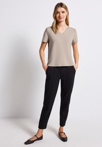 Béžový top s krátkým rukávem a V-výstřihem, kombinovaný s černými elegantními kalhotami. Modelka stojí se zhandy v kapsách a má na sobě černé baleríny.