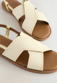 Beige Sandalen aus Kunstleder mit Kreuzriemen und einer braunen Gummisohle. Verfügt über einen goldenen Akzent am Fersenriemen und sichtbare Nähte.