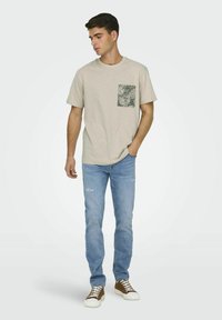 T-shirt beige con una tasca verde a motivo, abbinata a jeans chiari strappati e scarpe marroni con suole bianche. Look semplice e casual.
