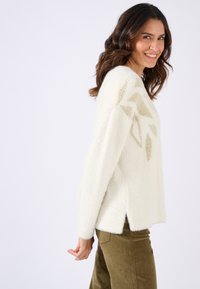 Suéter blanco y esponjoso que presenta un diseño texturizado de hojas en dorado claro. El suéter tiene una abertura lateral y se combina con pantalones verde oliva.