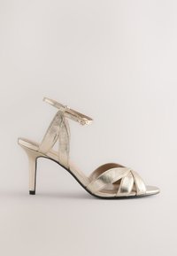 Sandalo con tacco metallizzato oro con cinturini incrociati, cinturino alla caviglia e suola nera elegante. Design a punta aperta, texture liscia.