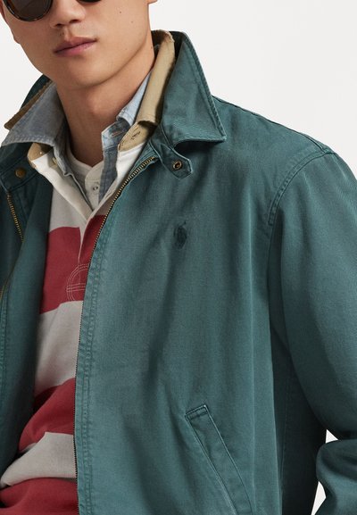 Siauros žalios spalvos lengva striukė su apykakle, užsegama užtrauktuku, su šoninėmis kišenėmis ir mažu logotipu. Dėvima ant raudonos ir baltos dryžuotos marškinėlių.