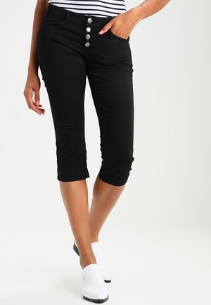 Femme portant un pantalon court noir avec quatre boutons argentés et des chaussures blanches sans lacets, main reposant dans la poche.
