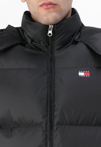 Melnas pūkainās jakas ar augstu apkakli, priekšējo rāvējslēdzēju un lieliem polsterētajiem iedaļām siltumizolācijai. Iezīmēta ar mazu Tommy Jeans logo.