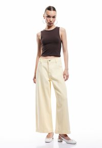 Haut sans manches marron associé à un jean jaune à taille haute et pattes d'éléphant. Le mannequin porte des chaussures à talons bloc blanches. Design simple et épuré.