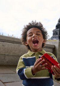Laps, kes kannab rohelist ja sinist triibulist kampsunit, hoiab punast mänguautobussi, naeratades laialt. Taustal on kivist arhitektuur ja pilvine taevas.