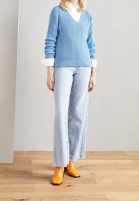 Maglione a coste blu chiaro sopra una camicia bianca, abbinato a jeans larghi blu chiaro e stivaletti con tacco arancioni brillanti.