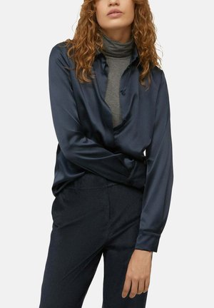 Vrouw draagt marineblauw satijnen overhemd over een grijze coltrui en donkere broek, poserend met één arm gekruist en de andere hangend.