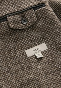 Braun texturiertes Gewebe mit einem gewebten Muster, ausgestattet mit einem Knopfverschluss, einer Brusttasche und einem Etikett mit der Aufschrift "NEXT REGULAR FIT."
