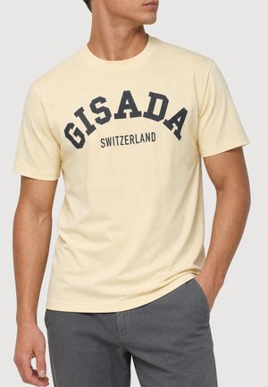 Mann trägt ein hellgelbes kurzärmeliges T-Shirt mit dem Text "GISADA Switzerland" und graue Hosen, eine Hand in der Tasche, leicht nach rechts gewandt.