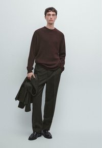 Brązowy sweter z dzianiny, ciemnoszare spodnie w paski i czarne buty; model trzyma pasującą kurtkę. Miękka tekstura, luźny krój.