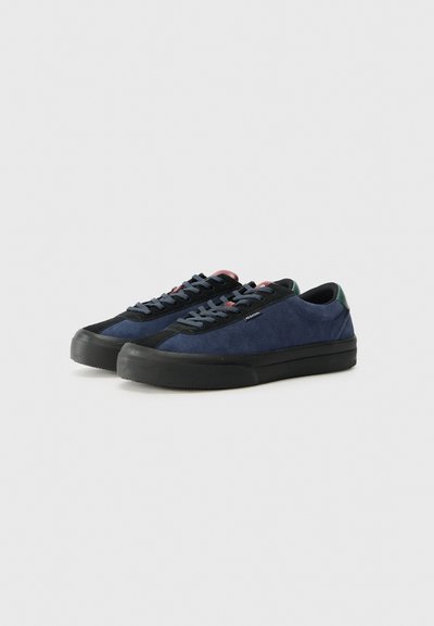 Adidași din suede navy cu talpă din cauciuc negru, cu sireturi închise la culoare și un accent verde în spate. Textură netedă cu un design cu profil jos.