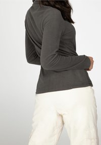 Kvinde iført en mørkegrå langærmet bluse og off-white bukser, der står let skråt vendt mod en ensfarvet hvid baggrund.