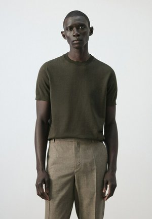 Hombre con cabello corto que lleva un suéter de manga corta verde oscuro y pantalones beige de talle alto con pliegues, contra un fondo claro y liso.