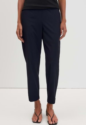 Person wearing black tapered pants and black heeled sandals, standing against a plain white background.