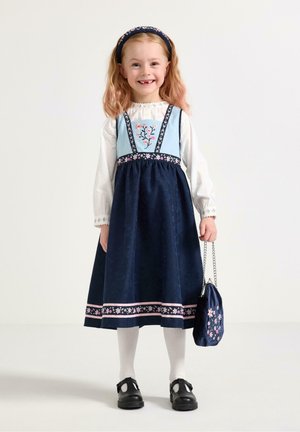 Smilende pige i mørkeblå kjole med blomsterbroderi, hvid bluse, hvide strømpebukser, sorte sko, blomsterhovedbøjle, holder en matchende taske.