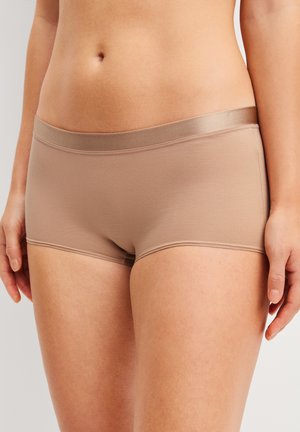 Frau trägt beige nahtlose Hipster-Unterwäsche, die den Mittelteil und die oberen Oberschenkel vor einem neutralen Hintergrund zeigt.