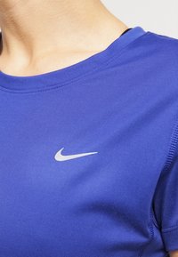 Nike sportovní tričko v jasně modré barvě, s kulatým výstřihem, krátkými rukávy a malým stříbrným logem na přední straně. Hladká textura.