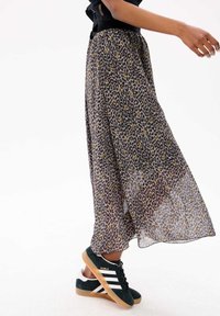 Jupe maxi transparente à imprimé léopard avec une taille noire, associée à des baskets Adidas Gazelle vertes avec des rayures blanches et une semelle en gomme.