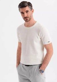 Suéter blanco de punto con mangas cortas y cuello redondo, con un patrón texturizado, combinado con pantalones gris claro y un reloj plateado.