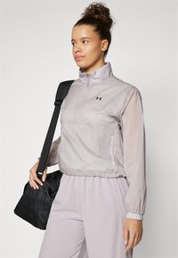 Lightweight, zip-up jacket in pale lavender, featuring long sleeves, elastic cuffs, and logo on the chest, paired with a black duffel bag.