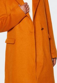 Abrigo de lana naranja con solapa con muesca, cierre de un botón, bolsillos laterales y una textura suave, mostrado parcialmente con una mano sosteniendo el cuello.