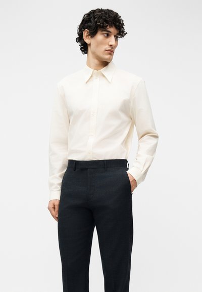 Άνδρας με σγουρά σκούρα μαλλιά που φοράει ανοιχτό μπεζ πουκάμισο με κουμπιά μακρυμάνικο και σκουρόχρωμο, καλοραμμένο παντελόνι, στέκεται με το ένα χέρι στην τσέπη.