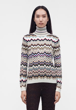 Femme portant un pull à col roulé à motif zigzag multicolore et un pantalon foncé à motif zigzag, debout devant un fond blanc uni.