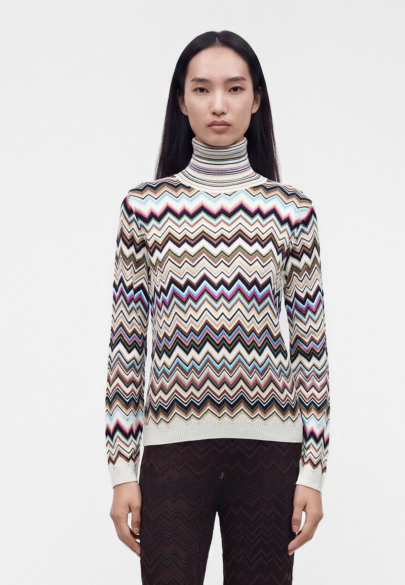 Femme portant un pull à col roulé à motif zigzag multicolore et un pantalon foncé à motif zigzag, debout devant un fond blanc uni.