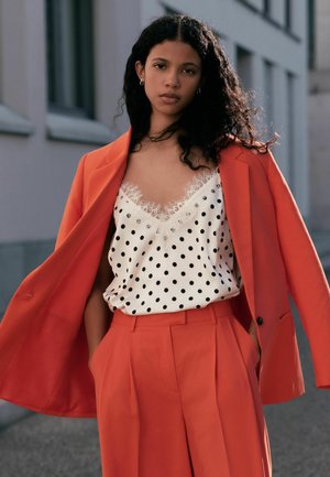 Ung kvinde med krøllet hår iført en orange dragt over en hvid polkaprikket top med blonder, stående udendørs med hænderne i lommerne.