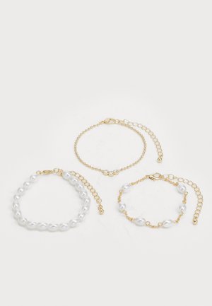 Tres pulseras: una con perlas blancas, una cadena dorada con acentos de cristal y una con eslabones mixtos de oro y perlas. Predominan los tonos dorados.